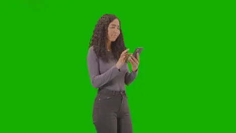 Three Quarter Length Shot Of Woman Waiting Looking At Mobile Phone Against Green Screen