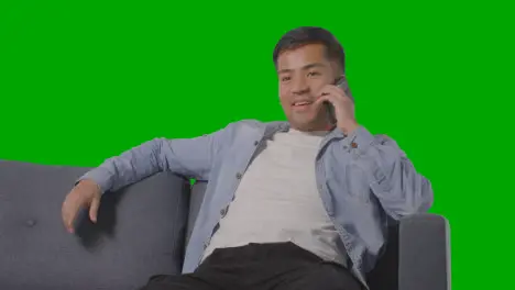 Studio Shot Of Young Man Sitting On Sofa Talking On Mobile Phone Against Green Screen 