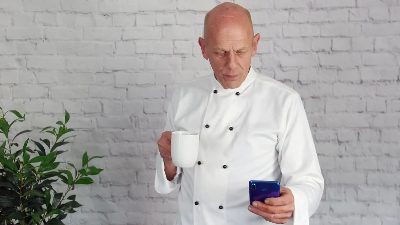 A mature senior head chef is taking a break from the kitchen and texting on his phone while drinking coffee