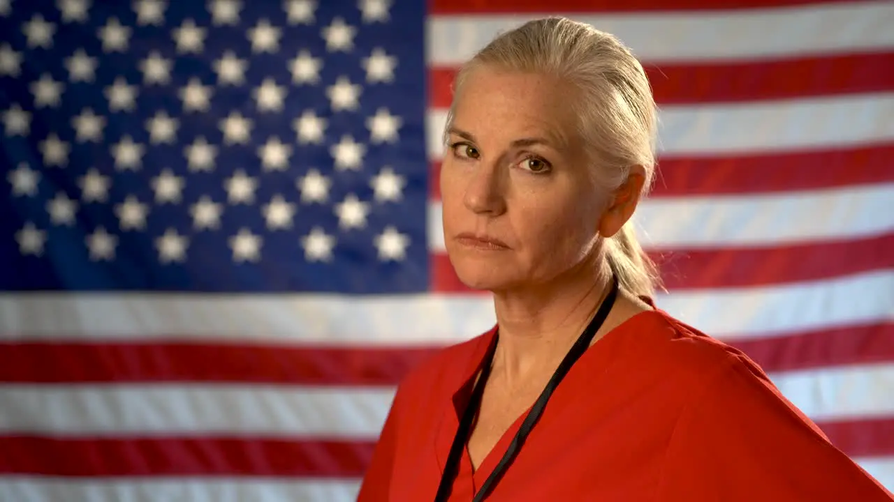 Medium tight portrait of the back of blonde nurses head as she turns quickly and looks angry and concerned with out of focus American flag