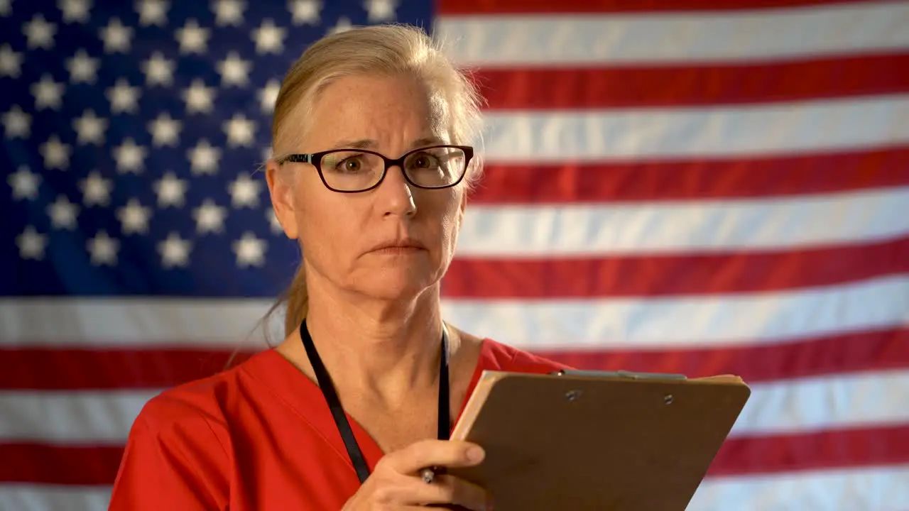 Medium tight portrait of concerned healthcare nurse looking at clipboard and then at camera with concerned look on her face with American flag behind her