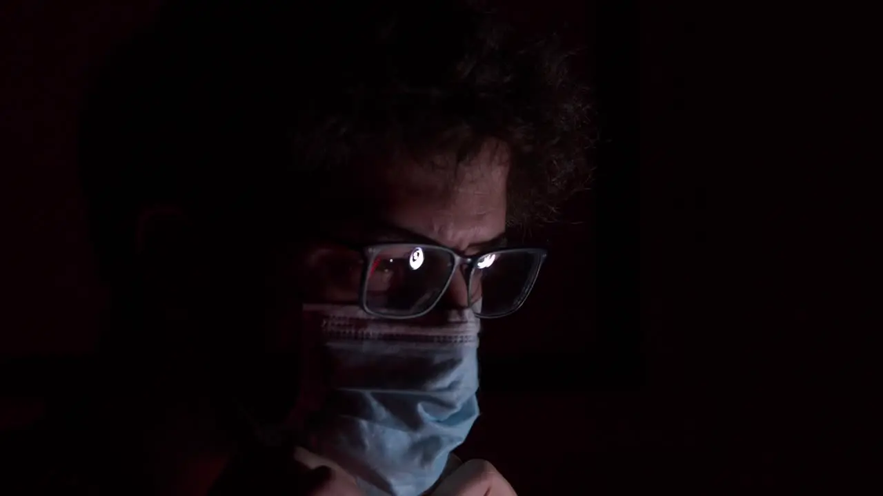 Young bearded male wearing glasses adjusting surgical corona virus mask in darkness