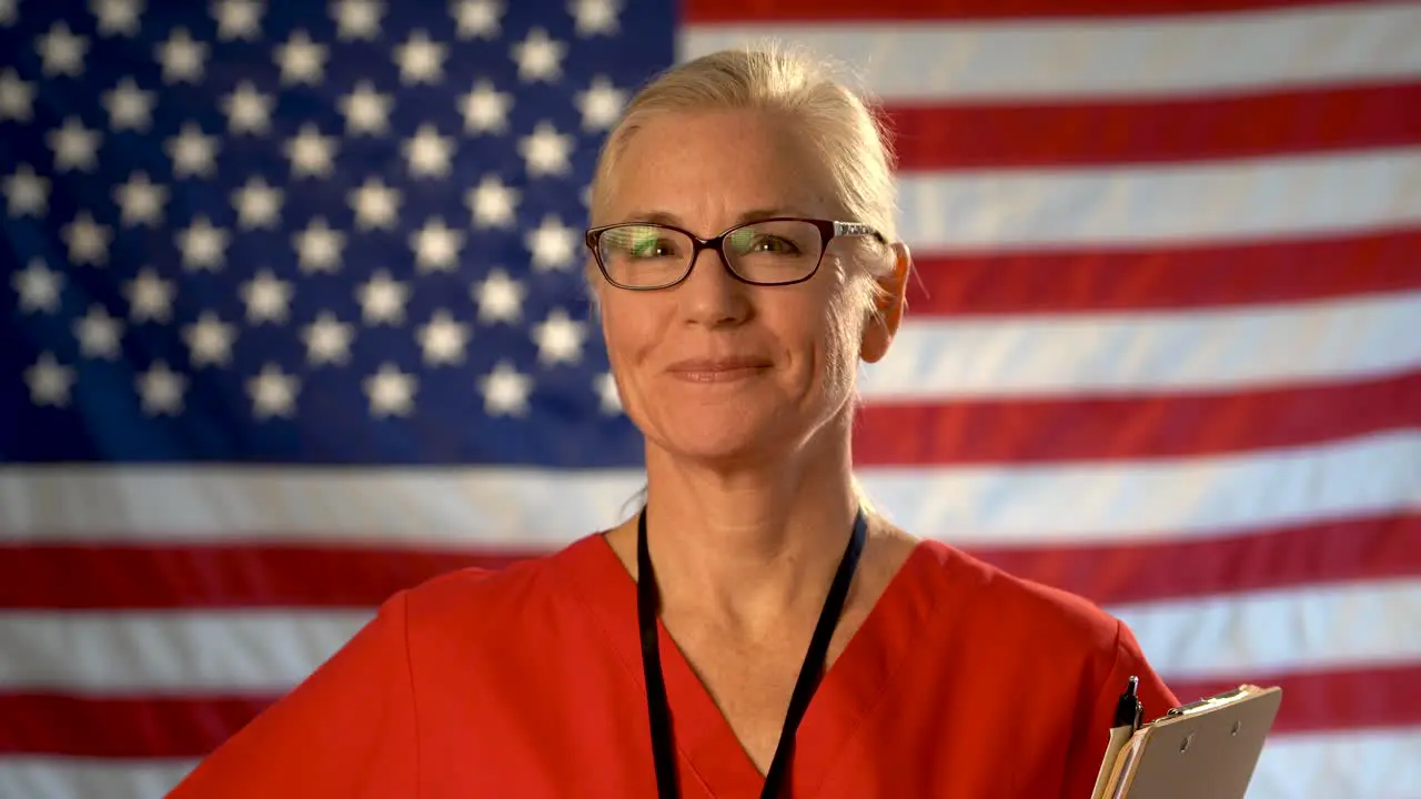 Medium tight portrait of a healthcare nurse with glasses walking toward camera happy with an out of focus American flag background