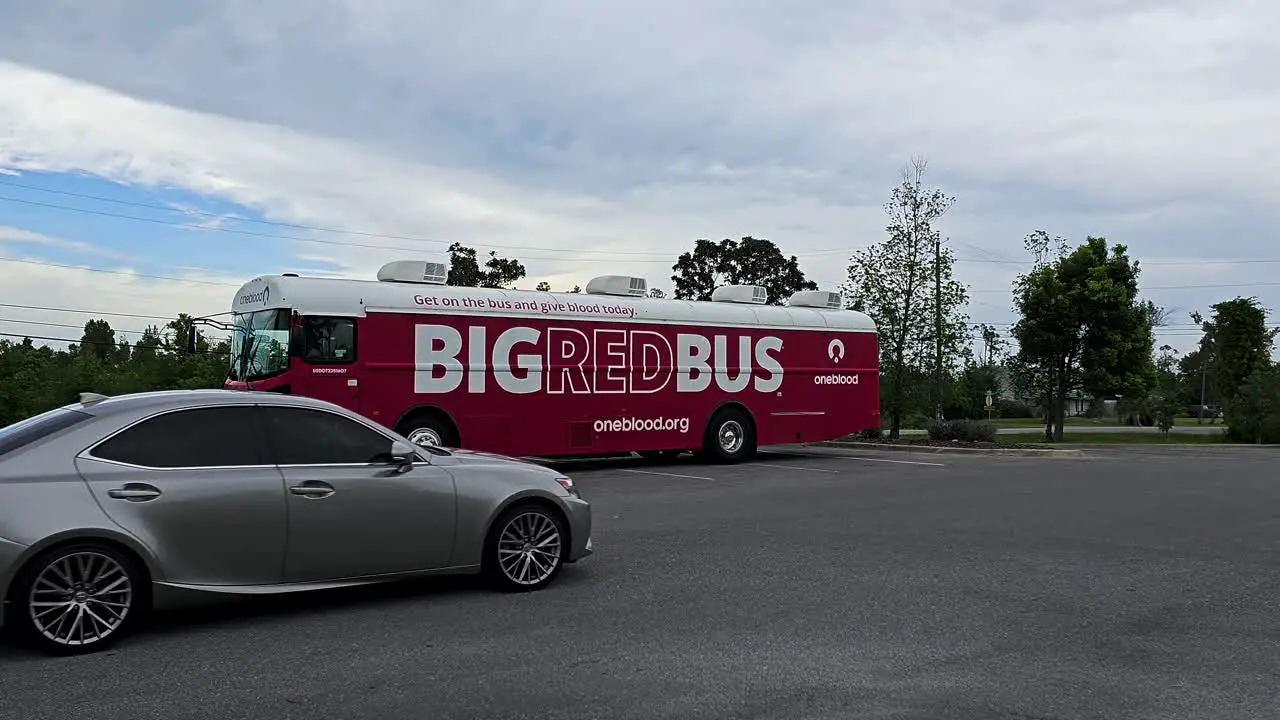 Oneblood big red bus blood donation vehicle sit in parking lot behind facility with silver car driving by