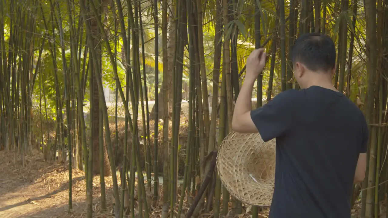 A man walking through a bamboo garden removes his hat to itch his scalp and brush the dandruff off his shirt