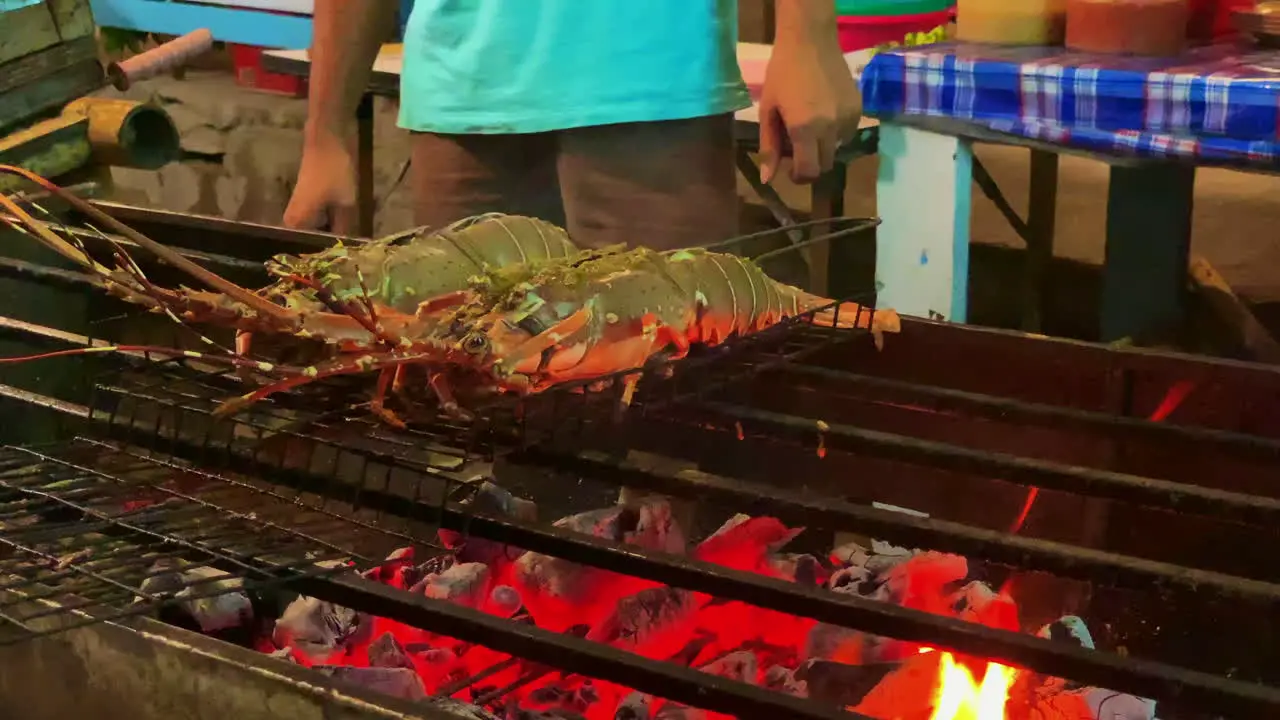 A close up shot of 2 lobsters while on the barbecue with flame metal grill on person unrecognisable in the background