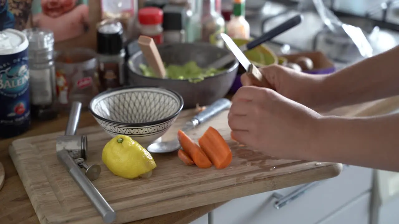 Hands of woman cutting carrot on cutting board next to lemon avocado halves and garlic press in a messy kitchen