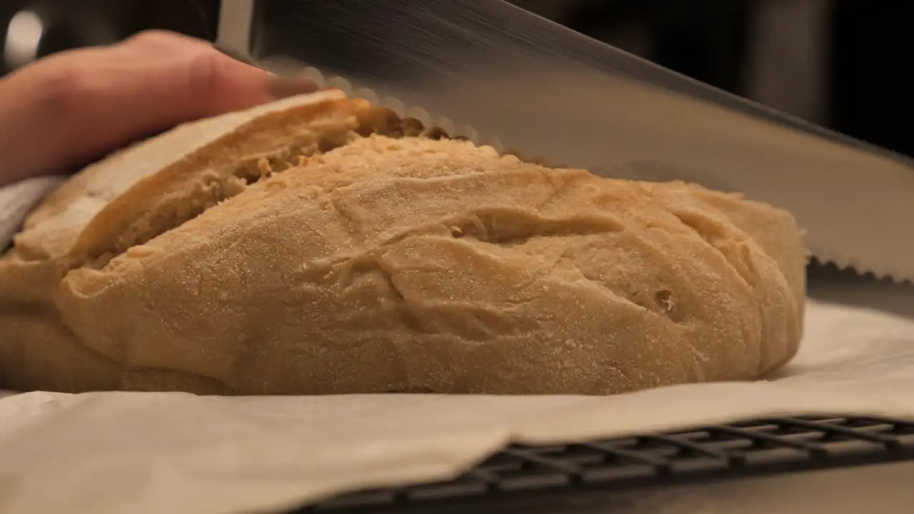 Person cutting through freshly baked loaf of sour dough bread with sharp serrated kitchen knife on bench top