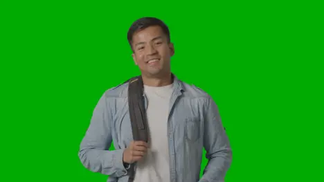 Portrait Of Smiling Male University Or College Student Putting On Backpack Ready For Class Against Green Screen 