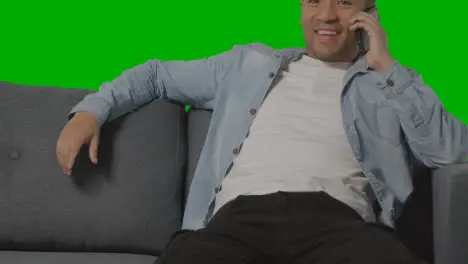 Close Up Of Young Man Sitting On Sofa Talking On Mobile Phone Against Green Screen 