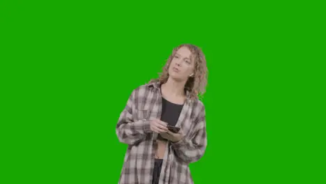 Studio Shot Of Young Woman Listening To Music On Mobile Phone And Dancing Against Green Screen