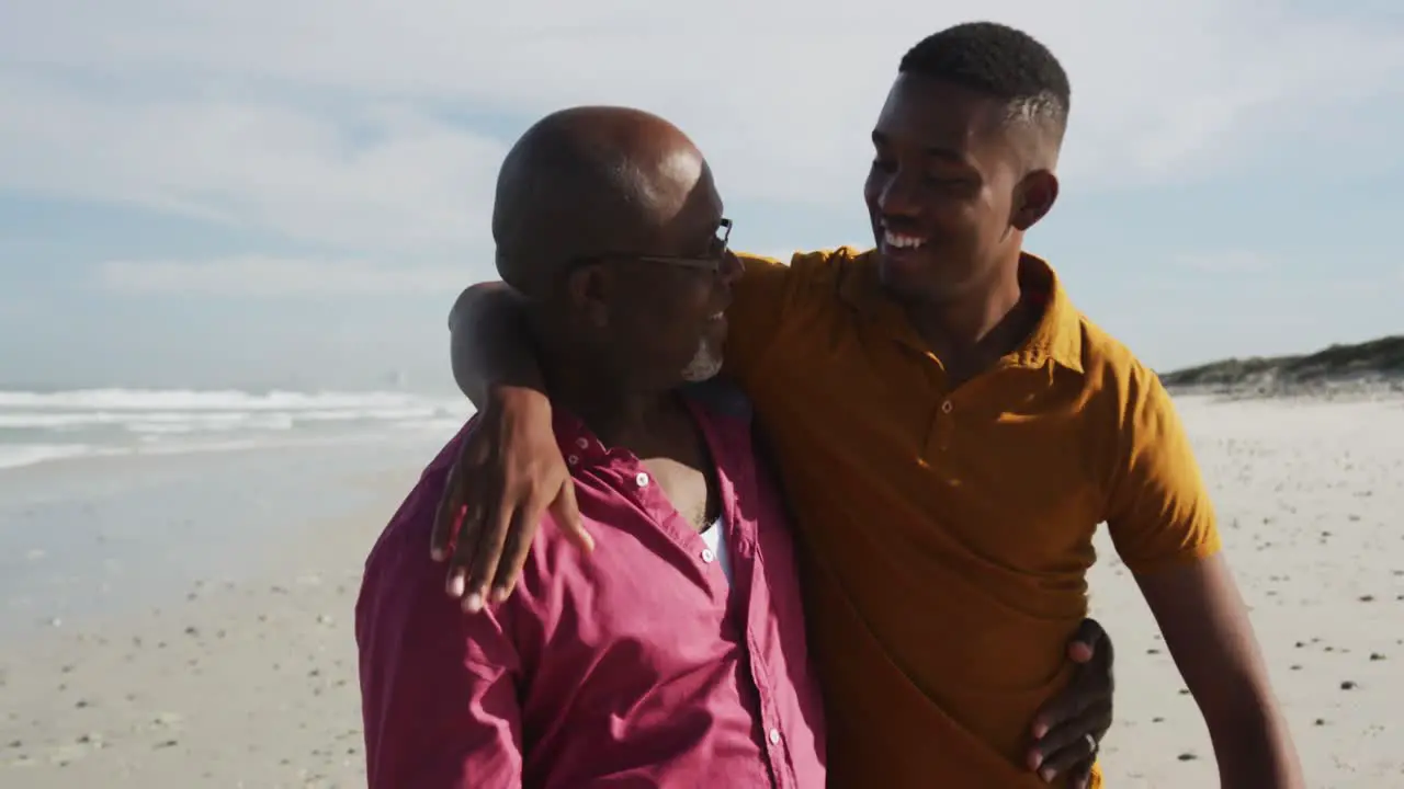 African american senior father and teenage son standing on beach embracing and talking