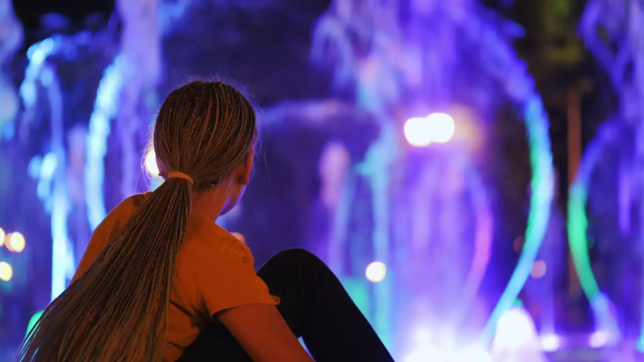 The child admires the fountain with lighting sits on the edge of the fountain rear view