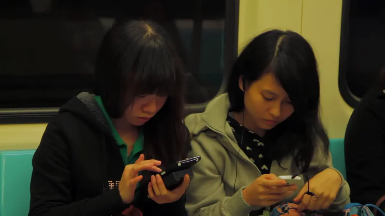 Two young Asian adult women busy with their phones in the subway