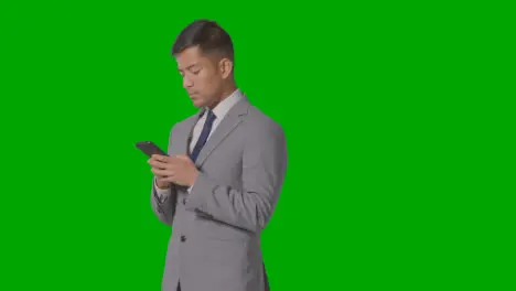 Studio Shot Of Businessman In Suit Looking At Mobile Phone Against Green Screen 