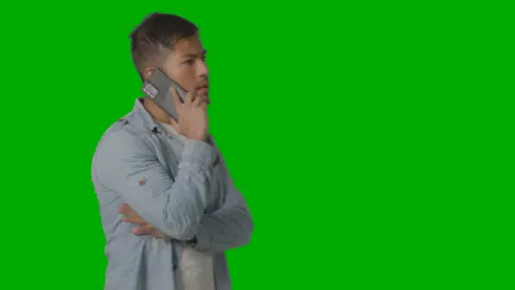 Studio Shot Of Casually Dressed Young Man Walking Across Frame Talking On Mobile Phone Against Green Screen 