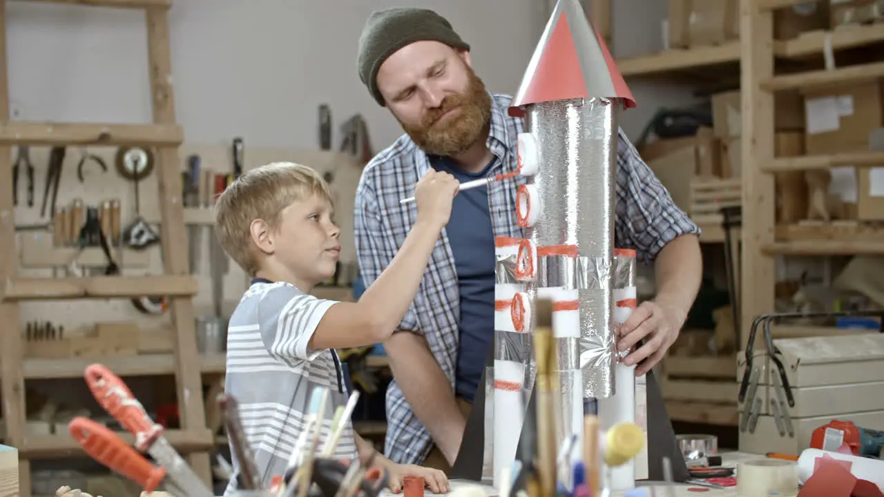 Kid Paints A Handmade Space Rocket With The Help Of His Father In The Craft Workshop