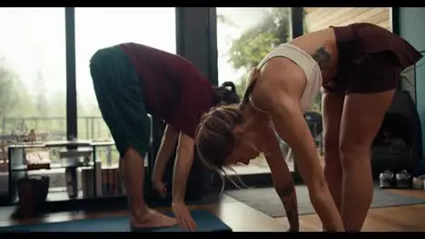 A brunette guy in a red T-shirt and a blonde girl in a white top are doing yoga and doing exercises in an industrial house overlooking a green forest