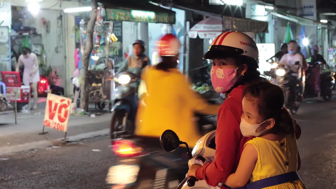 A mother and young daughter try to pull in to the steady stream of motor scooter traffic at night