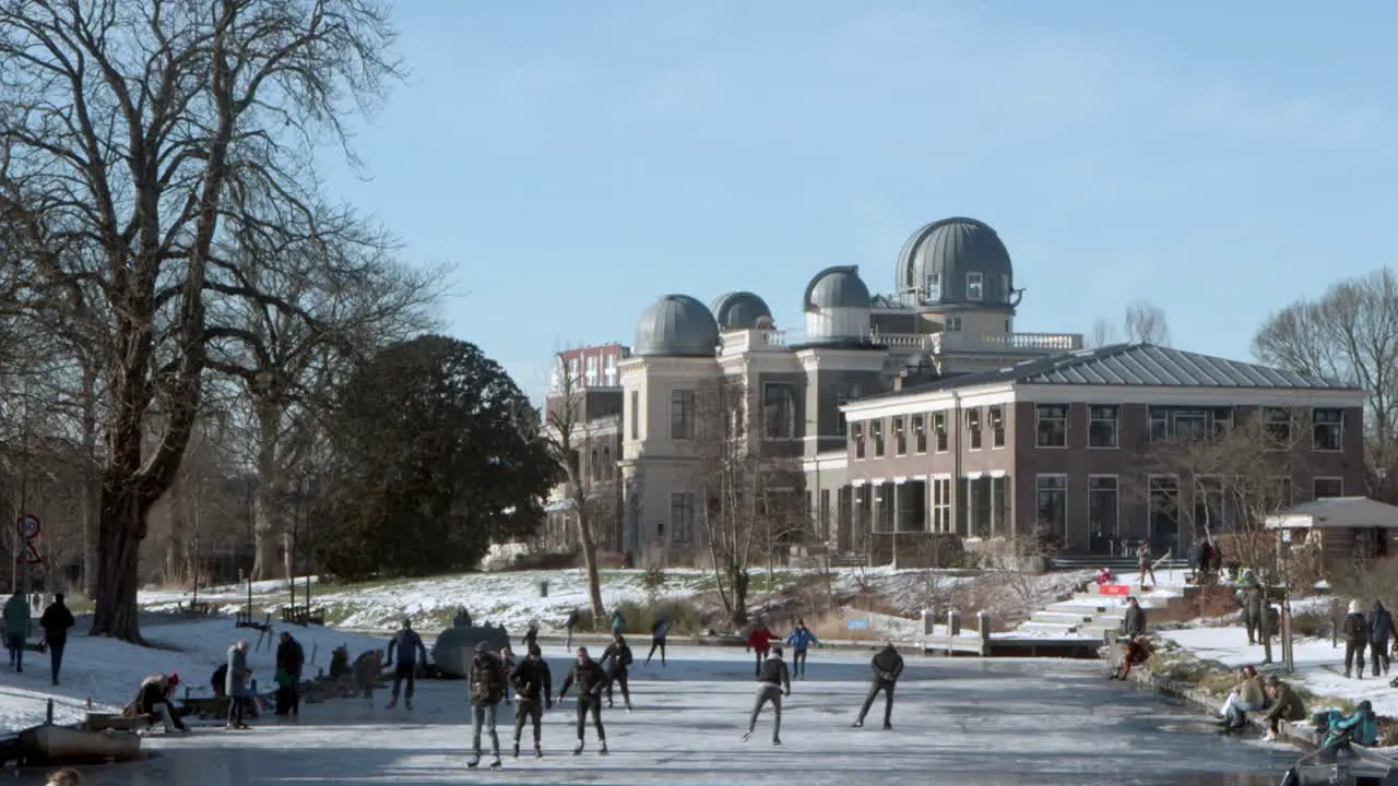 People ice skating on a frozen canal in front of the Leiden Observatory in the Netherlands on a sunny winter day during the pandemic