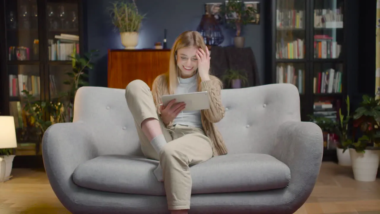 Happy Young Woman With Earphones And Tablet Entering Living Room And Sitting On Couch