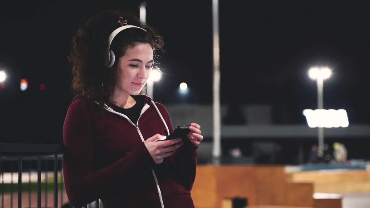 Smiling Sportswoman Listening Music With Bluetooth Headphones And Texting On Her Mobile Phone While Taking A Break During Her Training Session At Night In The Park