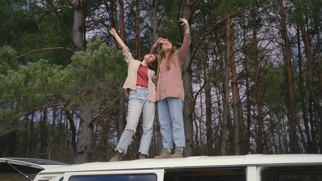 Two Young Girl Friends Take A Selfie With Each Other On Top Of The Roof Of A Caravan In The Middle Of The Forest