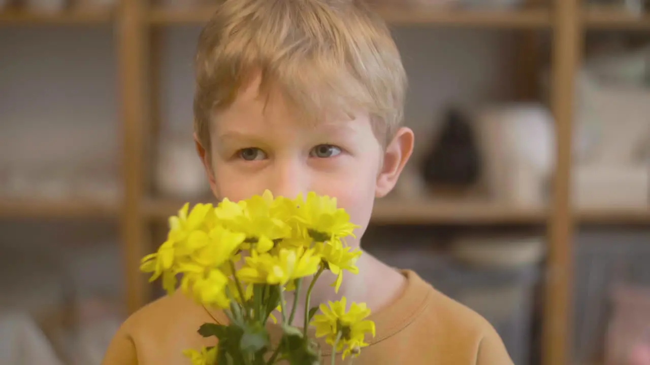 Close Up View Of A Blond Kid Smelling Yellow Flowers And Looking At Camera In A Craft Workshop