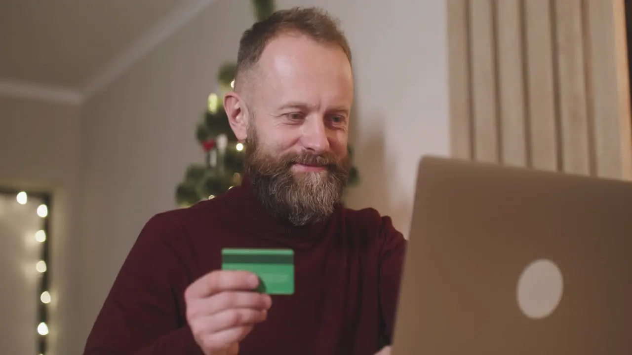 Vista Inferior De Un Hombre De Cabello Rojo Que Compra En Línea Con Una Tarjeta De Crédito Usando Una Computadora Portátil Sentada En Una Mesa En Una Habitación Decorada Con Un árbol De Navidad 1