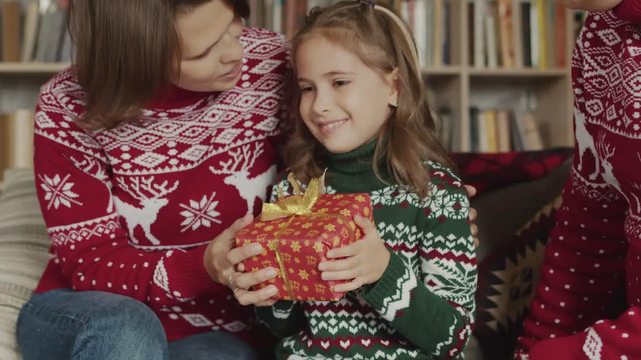 Linda Niña Sosteniendo Y Sacudiendo El Regalo De Navidad Mientras Se Sienta Con Sus Padres En El Sofá En Casa