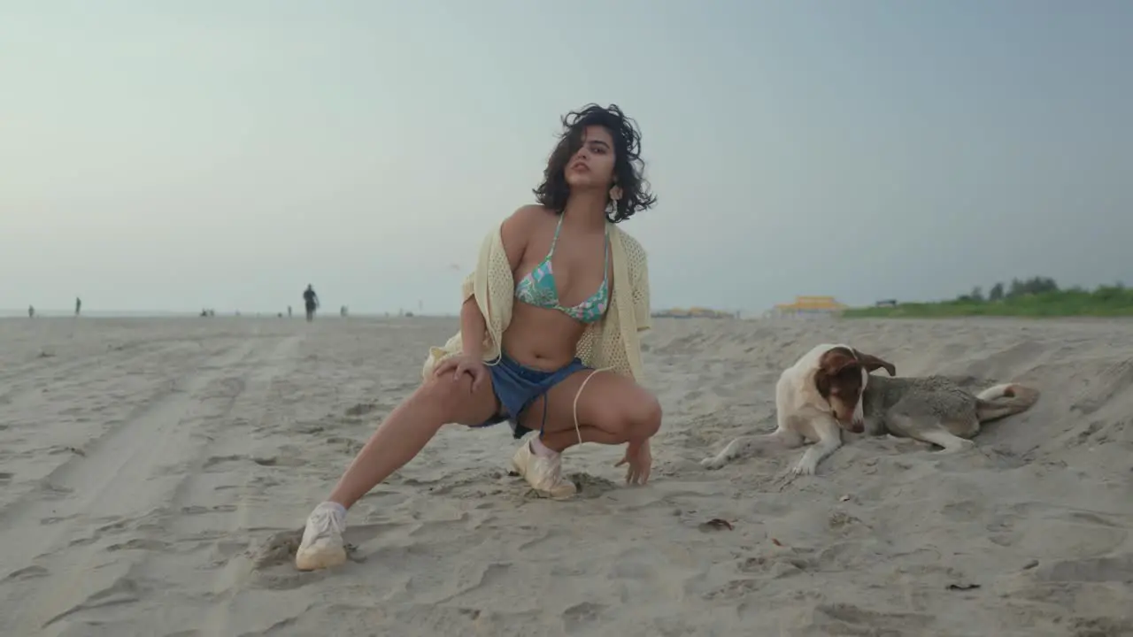 Young woman in a playful pose on a sandy beach with a dog lying next to her casual beachwear overcast day