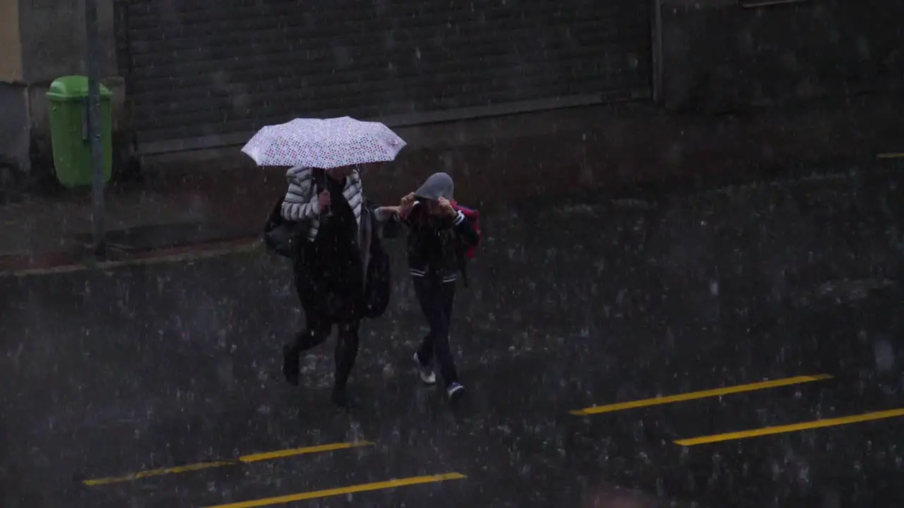 Mother and sunning crossing a street during a hail storm protecting themselves with an umbrella and a hood