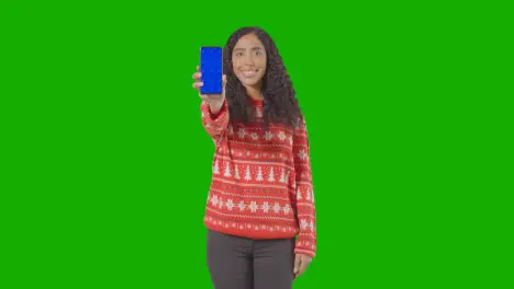 Studio Portrait Of Woman Wearing Christmas Jumper Holding Up Mobile Phone Against Green Screen