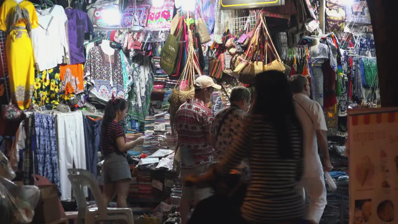 Medium Exterior Still Shot of Tourists Exploring Night Market Wile Seller Sorts Out Her Sock From Across a Road in the Night Time