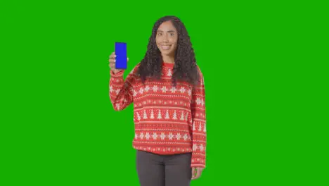 Studio Portrait Of Woman Wearing Christmas Jumper Holding Up Mobile Phone Against Green Screen 1