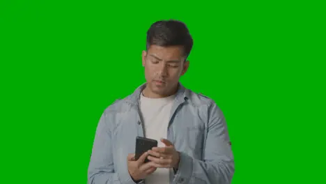 Studio Shot Of Casually Dressed Young Man Messaging On Mobile Phone Against Green Screen 