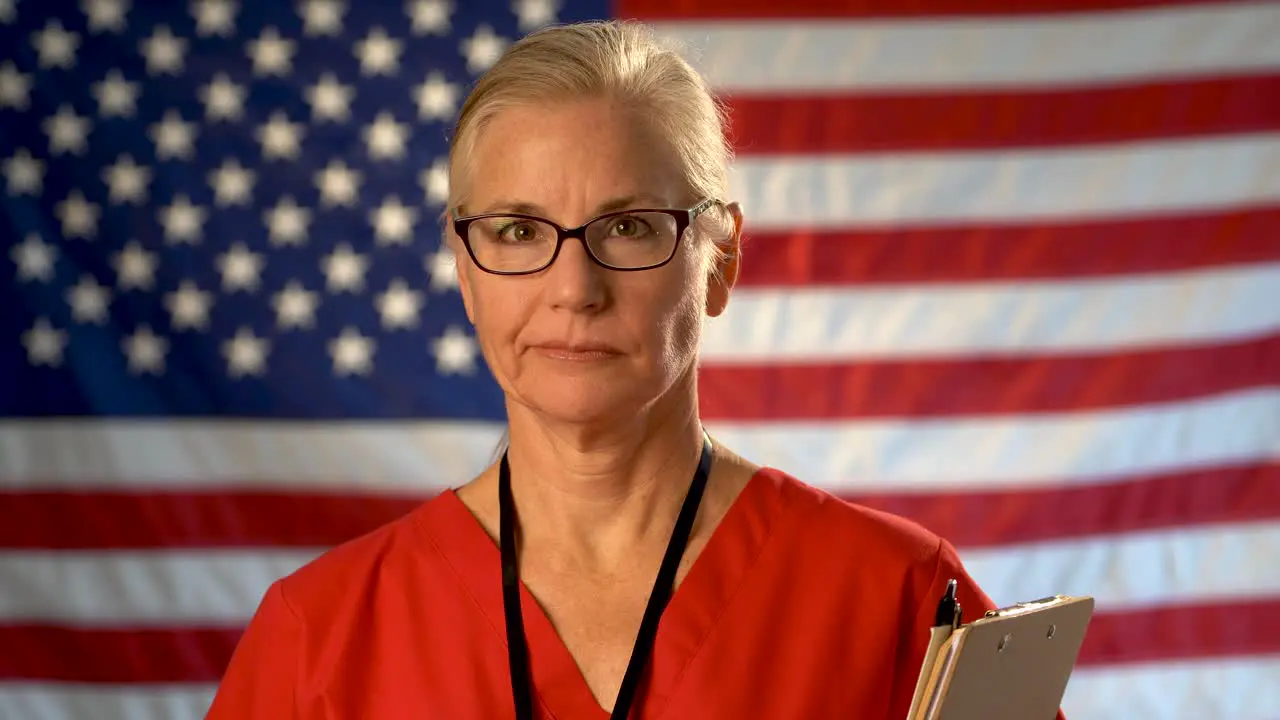 Medium tight portrait of a healthcare nurse with clipboard walking towards camera shaking her head with an out of focus American flag