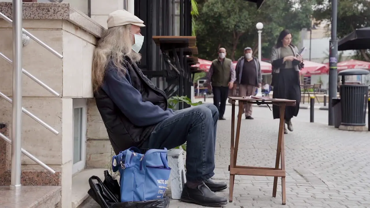 An elderly man in a face mask sitting on the edge of a sidewalk with his back to the street