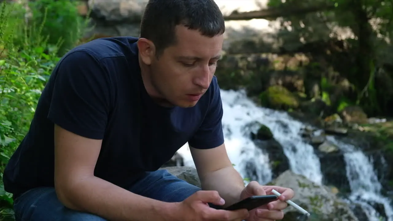 Young alone man browsing social media on a phone and smoking cigarette near a waterfall