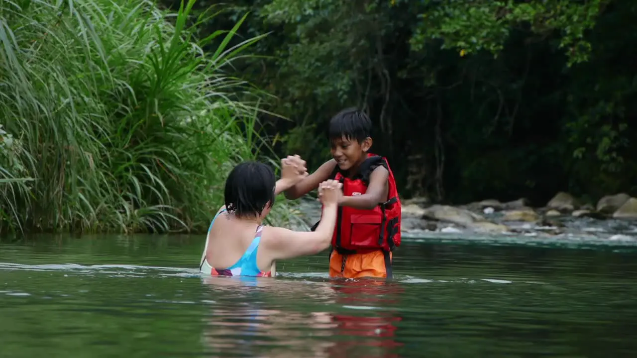 Indian boy wearing red life jacket playfully hand wrestling with Asian woman in river