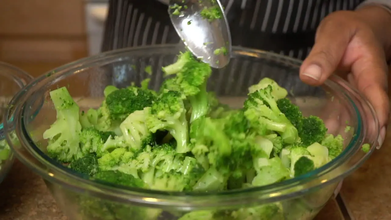 Cook stirs steamed broccoli in glass bowl with tablespoon slow motion