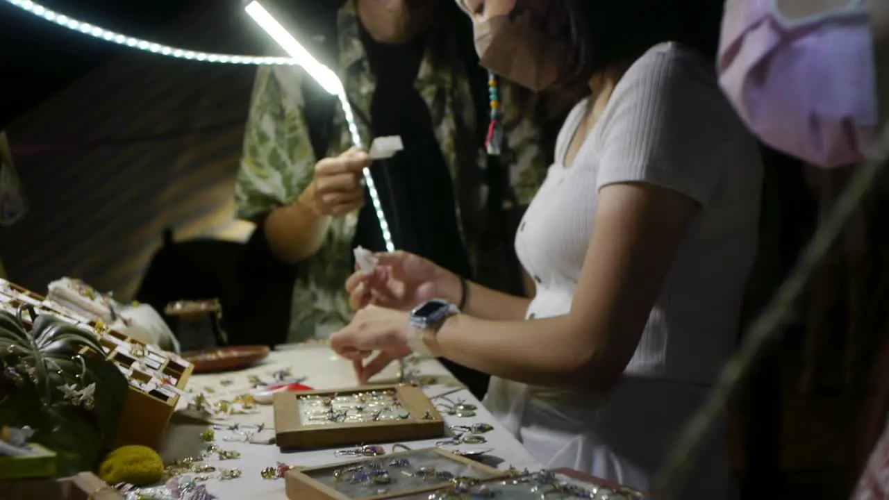 Asian woman wearing face mask while browsing jewelry at outdoor night market filmed handheld with slow downward tilt