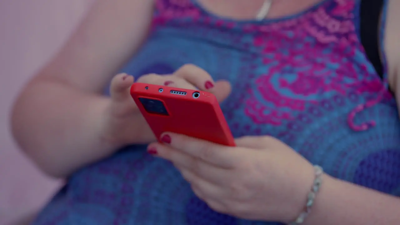 Close up of a fat woman scrolling and playing with her red Smartphone while sitting wearing a colourful blue pink dress and red nail polish in slow motion