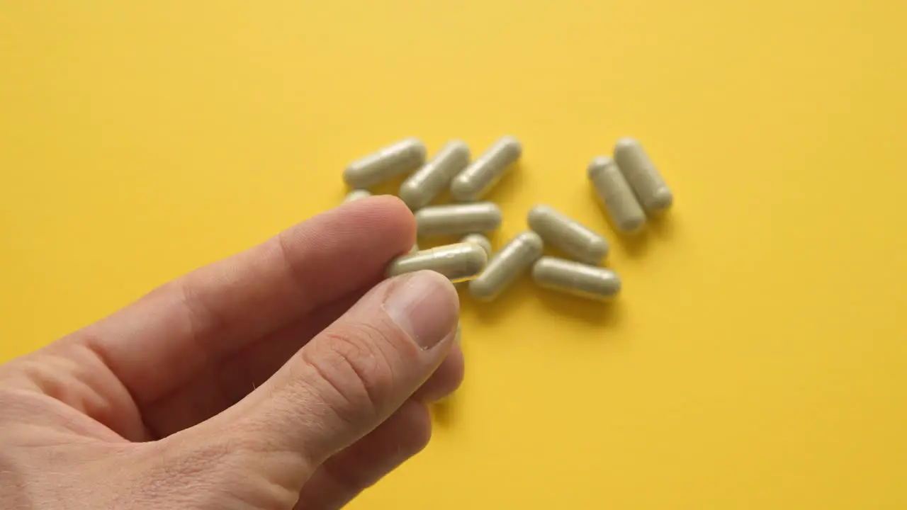 Left Male Hand Holding Lions Mane Supplement Pill In Between Fingers With Yellow Background With Pills On Table