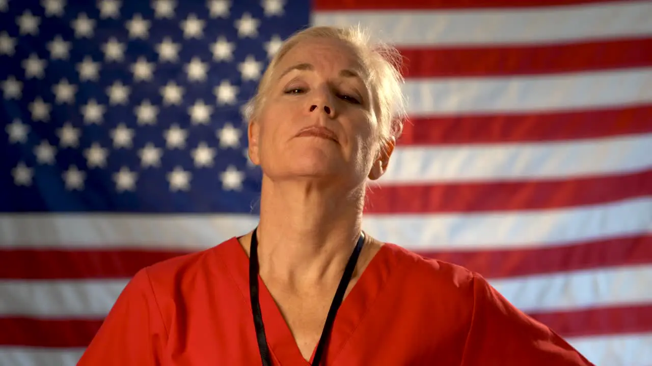 Medium tight portrait of a healthcare nurse taking glasses off and looking concerned and sympathetic with an out of focus American flag