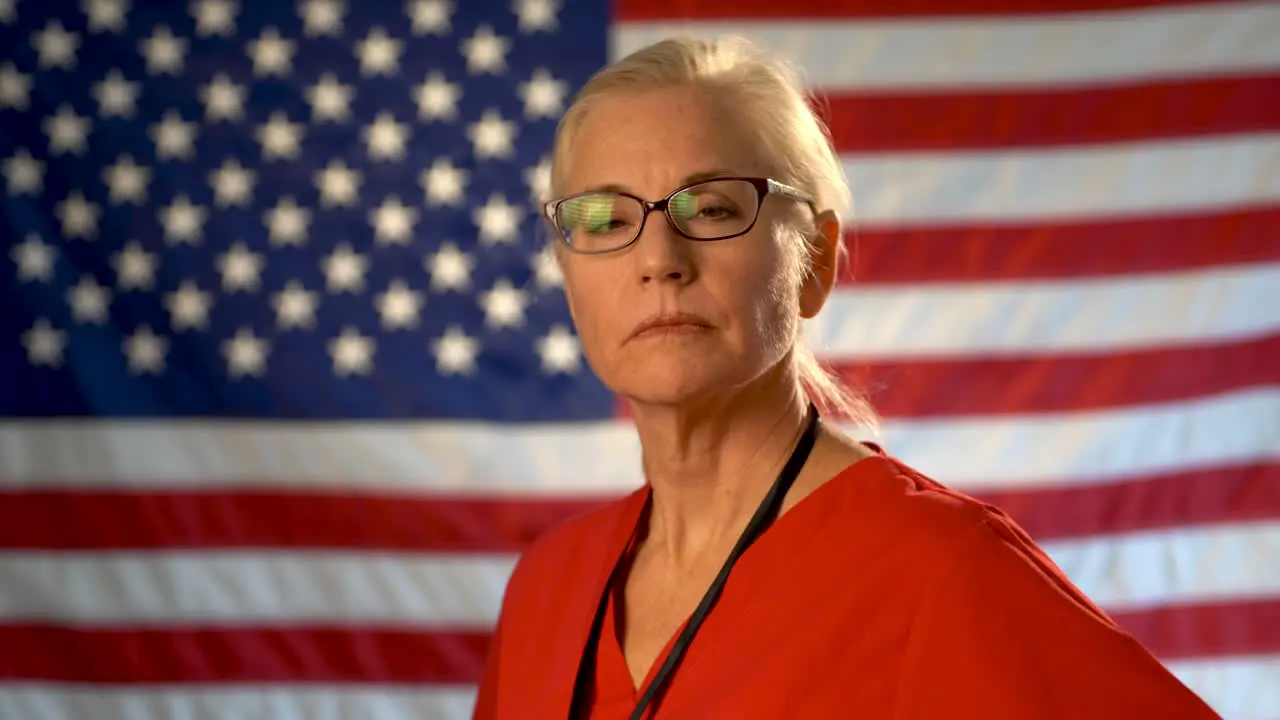 Medium tight portrait of the back of blonde nurses head as she turns and looks at camera looking angry wearing glasses with out of focus US flag