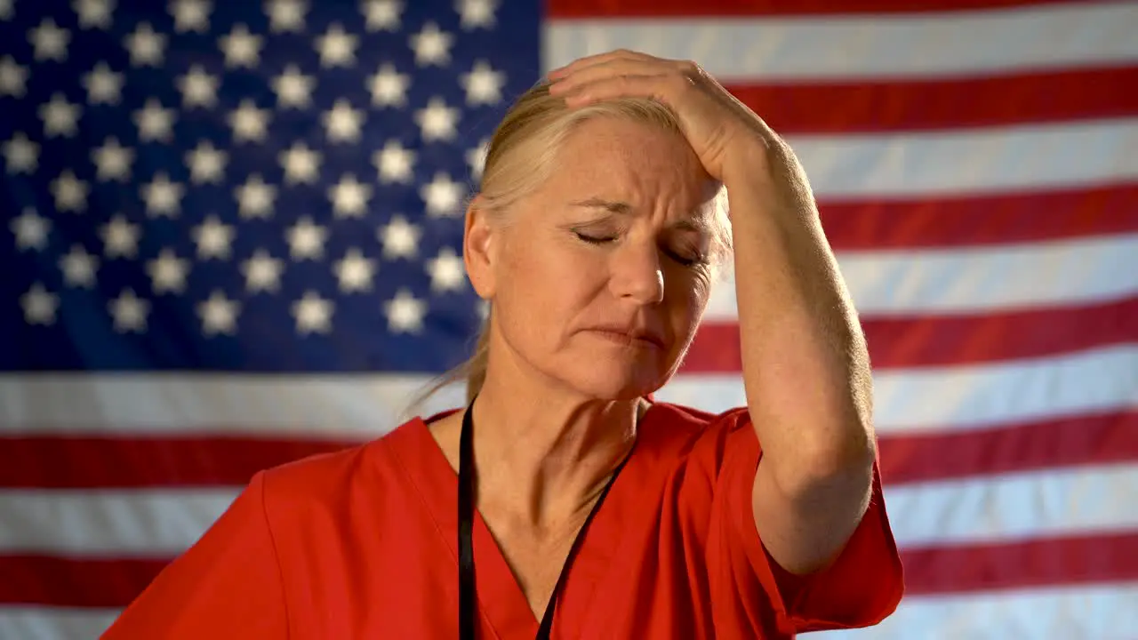 Medium tight portrait of nurse looking very worried and sad rubbing her forehead with American flag behind her