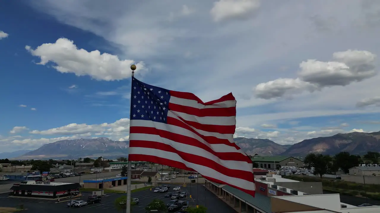 American flag USA blowing waving in the wind on beautiful sunny summer day with clouds and blue skies overlooking mountains and small town America USA as drone flys pans around flagpole in 4K 60fps