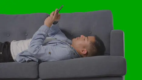 Studio Shot Of Man Lying On Sofa Scrolling Through Mobile Phone Against Green Screen