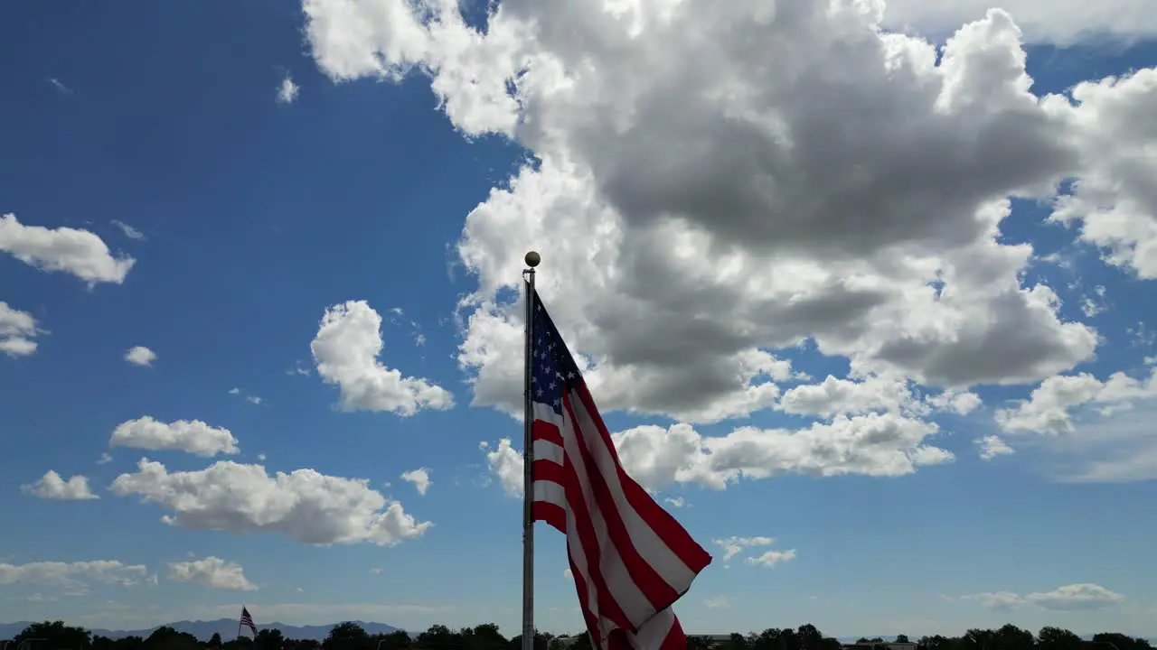 American flag USA blowing waving in the wind on beautiful sunny summer day with clouds and blue skies as drone flys around flagpole with small town cresting in bottom of shot in 4K 60fps